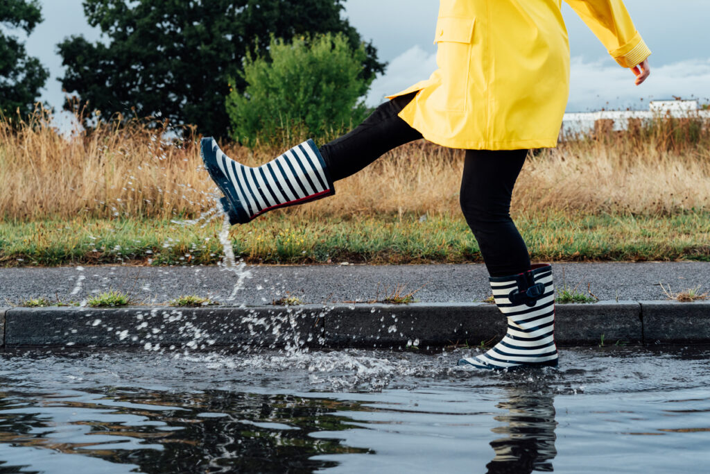 Woman having fun on the street after the rain. Cropped woman wearing rain rubber boots and yellow raincoat walking into puddle with water splash and drops. Fall weather. Selective focus.